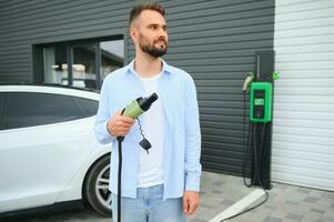 Man Holding Power Charging Cable For Electric Car In Outdoor Car Park. And he s going to connect the car to the charging station in the parking lot near the shopping center. photo