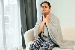 Young Muslim man suffering from a sore throat, sitting on the sofa at home and grimacing, holding his throat. Needs medical treatment photo