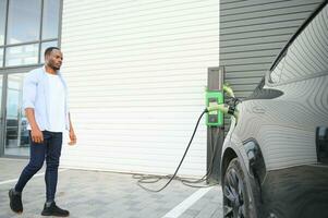 African man standing near electric car with charging cable in plug. Eco friendly vehicle charging on station. LIfestyles concept photo