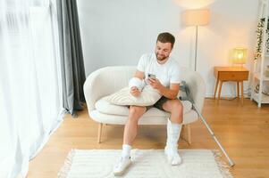 Man with a broken leg and arm using his mobile phone while relaxing on the sofa at home. Accident, injury, treatment, rehabilitation concept photo