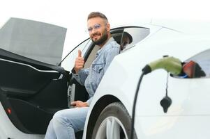 hermosa joven elegante hombre es con eléctrico coche a tiempo de día cargando el vehículo foto