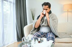 health, cold and people concept - sick young indian man in blanket having headache or fever at home photo
