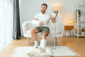 man recovery from accident fracture broken bone injury with leg splints in cast neck splints collar arm splints sling support arm in living room. Social security and health insurance concept photo