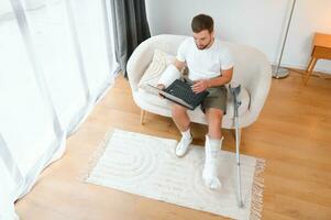 Unhealthy businessman with injured leg and arm in bandage sit on sofa at home work online on computer photo