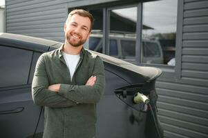 handsome bearded man in casual wear, standing at the charging station and charger for an electric car. Eco electric car concept photo