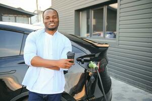 An African-American man is standing near an electric car, waiting for it to charge at a charging station, and drinking coffee. photo