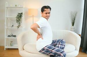 A sick man on Christmas has severe back pain, massages muscle his hand sitting on sofa in living room photo