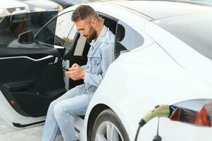 Beautiful young stylish man is with electric car at daytime charging the vehicle photo