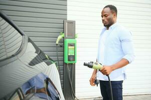 Serious african man holding charge cable in on hand, standing near luxury electric car. photo