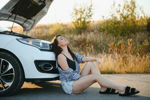 A young girl sits near a broken car on the road with an open hood. photo