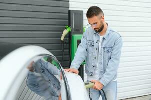 Beautiful young stylish man is with electric car at daytime charging the vehicle photo