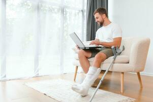 Happy young man with arm in a cast sitting on the couch at home and communicating on a laptop photo