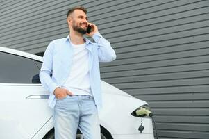 casual hombre con teléfono inteligente cerca eléctrico coche esperando para el terminar de el batería cargando proceso foto