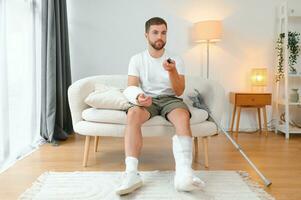 man recovery from accident fracture broken bone injury with leg splints in cast neck splints collar arm splints sling support arm in living room. Social security and health insurance concept photo