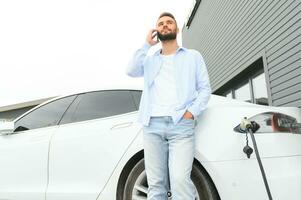 Happy man using smart phone and charging car at vehicle charging station photo