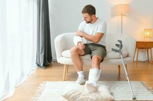 man recovery from accident fracture broken bone injury with leg splints in cast neck splints collar arm splints sling support arm in living room. Social security and health insurance concept photo