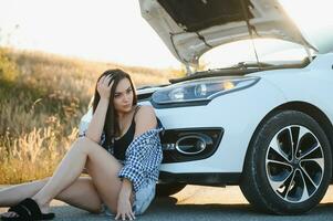 hermosa joven niña sentado a un roto coche en el la carretera desesperado a obtener ayuda foto