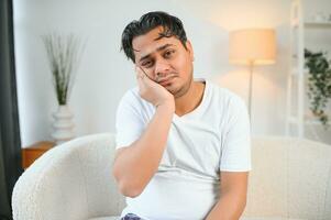 Dental problems. Young indian man touching cheek, closing eyes with expression of terrible suffer from painful toothache, sensitive teeth, cavities. Hindu guy at home apartment living room on sofa photo