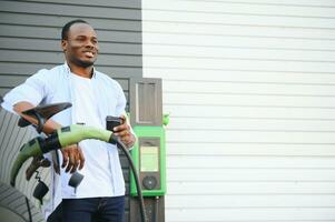 Stylish african man with coffe cup in hand inserts plug into the electric car charging socket photo