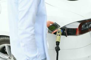 Happy man using smart phone and charging car at vehicle charging station photo