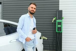 Happy man using smart phone and charging car at vehicle charging station photo