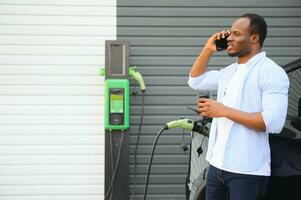 un africano hombre es en pie cerca un eléctrico auto, esperando para eso a cargar a un cargando estación y utilizando el teléfono. foto