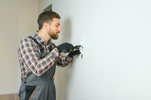 Confident professional electrician in uniform using screwdriver while replacing a socket in apartment after renovation work. photo