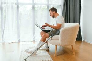 Happy young man with arm in a cast sitting on the couch at home and communicating on a laptop photo