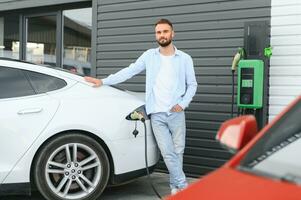 Beautiful young stylish man is with electric car at daytime charging the vehicle photo