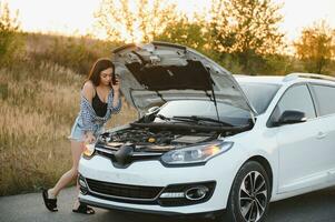 Attractive slim young girl in summer shorts and shirt repairs a broken car. A beautiful woman stands near raised car hood. photo