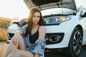 A young girl sits near a broken car on the road with an open hood. photo