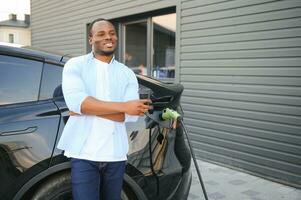 africano hombre en pie cerca eléctrico coche con cargando cable en enchufar. eco simpático vehículo cargando en estación. estilos de vida concepto foto