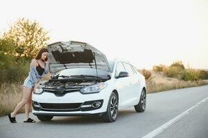 Beautiful slim girl in shirt and shorts looks in open car hood on a road photo