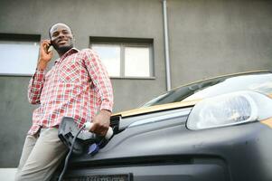 Electric car charging concept. portrait of young handsome dark skinned man, standing at the charging station and talking phone whie waiting for his car charging photo