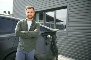 handsome bearded man in casual wear, standing at the charging station and charger for an electric car. Eco electric car concept photo