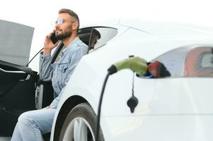 hermosa joven elegante hombre es con eléctrico coche a tiempo de día cargando el vehículo foto