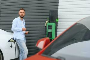 hermosa joven elegante hombre es con eléctrico coche a tiempo de día cargando el vehículo foto