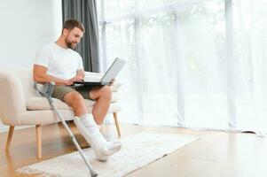 Unhealthy businessman with injured leg and arm in bandage sit on sofa at home work online on computer photo