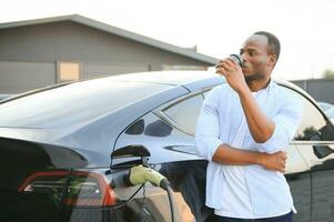 An African-American man is standing near an electric car, waiting for it to charge at a charging station, and drinking coffee. photo