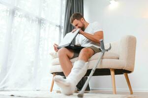 Unhealthy businessman with injured leg and arm in bandage sit on sofa at home work online on computer photo