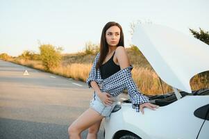 atractivo Delgado joven niña en verano pantalones cortos y camisa refacción un roto coche. un hermosa mujer soportes cerca elevado coche capucha. foto