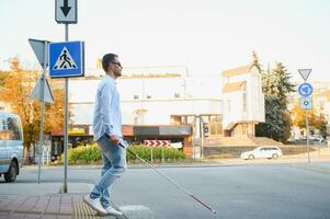 joven ciego hombre con blanco caña caminando a través de el calle en ciudad foto