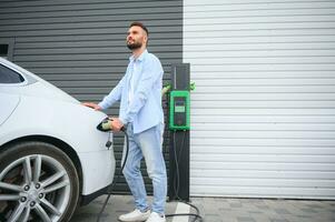 Beautiful young stylish man is with electric car at daytime charging the vehicle photo