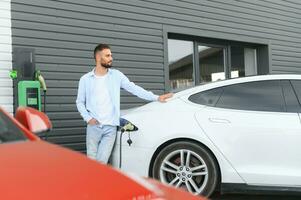 Beautiful young stylish man is with electric car at daytime charging the vehicle photo