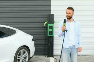 young handsome man holding charging cable at electric charging station point standing near his new car photo