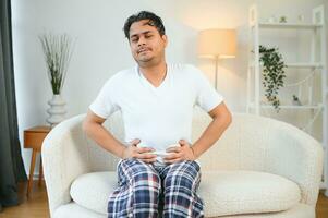 Unhappy indian or arabian man, sits on comfortable sofa in cozy living room, holds his hands on his stomach, grimaces from pain in his stomach, suffers from poisoning, spasm, stomach problems. photo