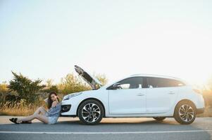 A young girl sits near a broken car on the road with an open hood. photo