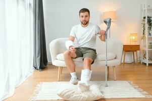 man recovery from accident fracture broken bone injury with leg splints in cast neck splints collar arm splints sling support arm in living room. Social security and health insurance concept photo