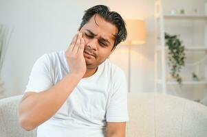 dental problemas. joven indio hombre conmovedor mejilla, clausura ojos con expresión de terrible sufrir desde doloroso dolor de muelas, sensible dientes, cavidades hindú chico a hogar Departamento vivo habitación en sofá foto