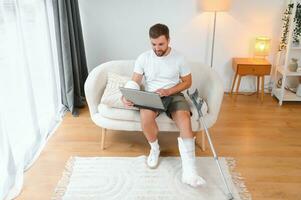 Unhealthy businessman with injured leg and arm in bandage sit on sofa at home work online on computer photo